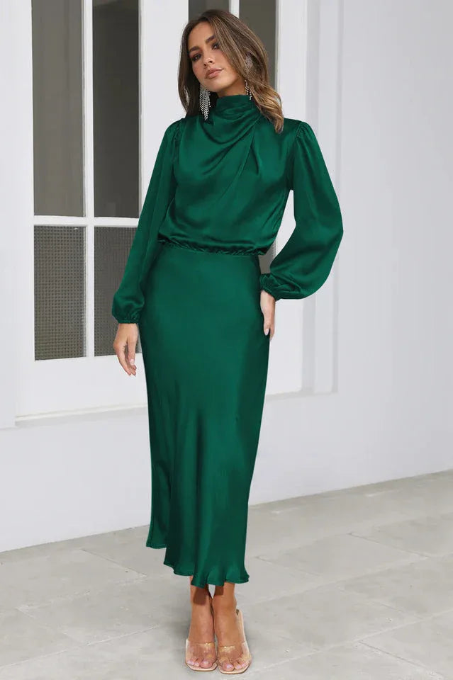 Woman wearing a long-sleeved emerald green satin dress with a cowl neckline and a ruffled hem, standing in a studio.