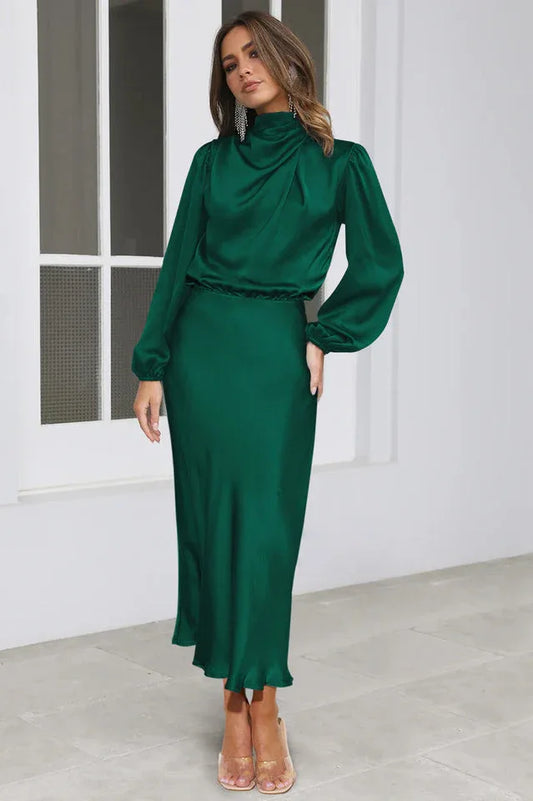 Woman wearing a long-sleeved emerald green satin dress with a cowl neckline and a ruffled hem, standing in a studio.
