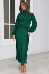 Woman wearing a long-sleeved emerald green satin dress with a cowl neckline and a ruffled hem, standing in a studio.