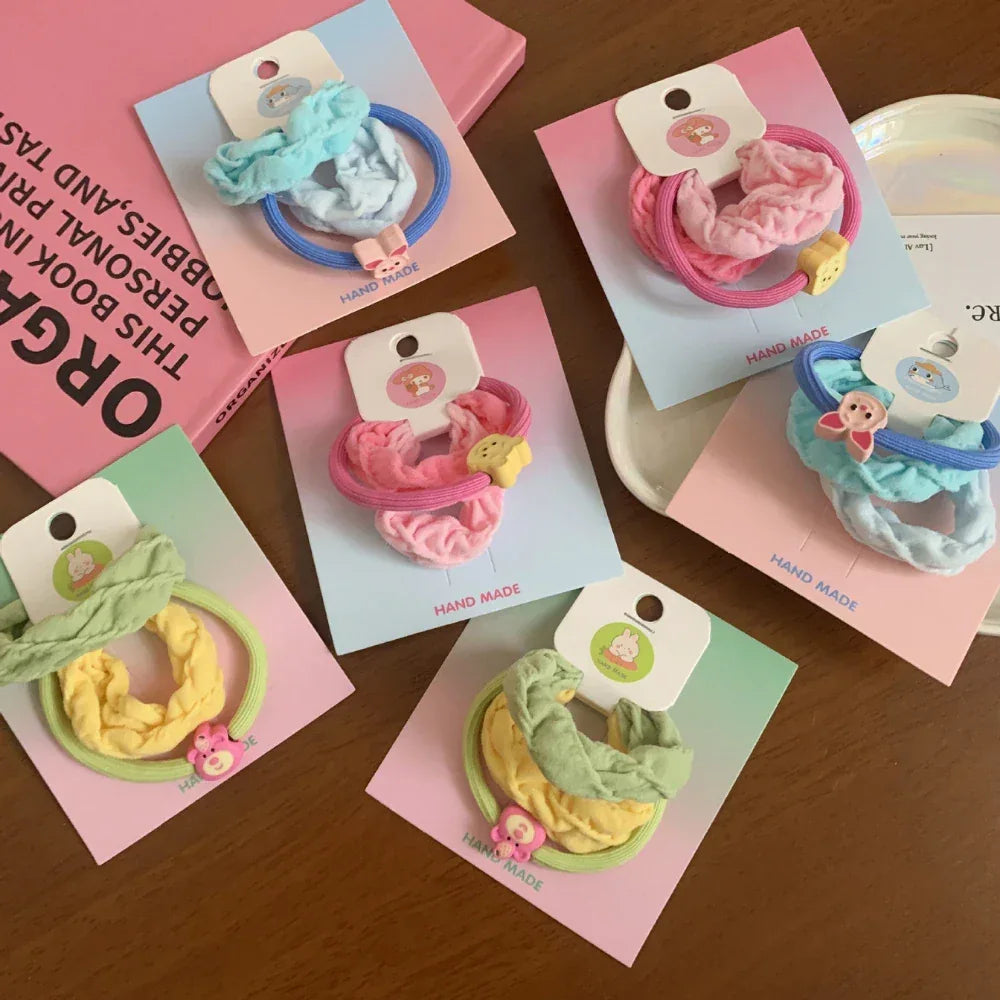 Several scrunchie sets with fabric ties in shades of blue, pink, yellow, and green, adorned with small animal charms, laid out on pastel backing cards.