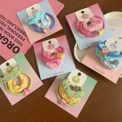 Several scrunchie sets with fabric ties in shades of blue, pink, yellow, and green, adorned with small animal charms, laid out on pastel backing cards.