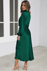 Back view of a woman wearing a long-sleeved emerald green satin dress with a cowl neckline and a ruffled hem, standing in a studio.
