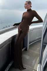 Woman wearing a long-sleeved, brown, mock neck, floor-length dress with a draped knot detail at the waist, posing on a balcony with a city view.
