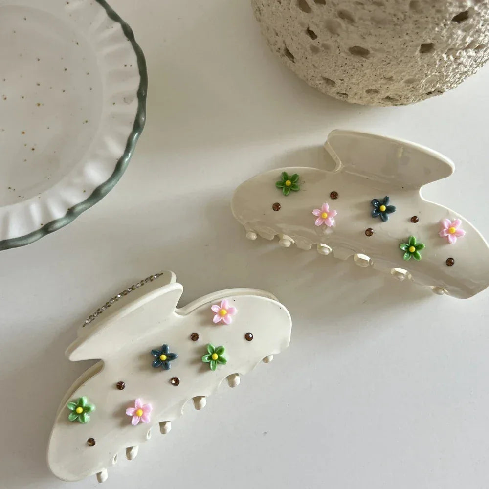 Two cream-colored claw clips with floral and gemstone embellishments, arranged on a white surface with a decorative plate and textured object.