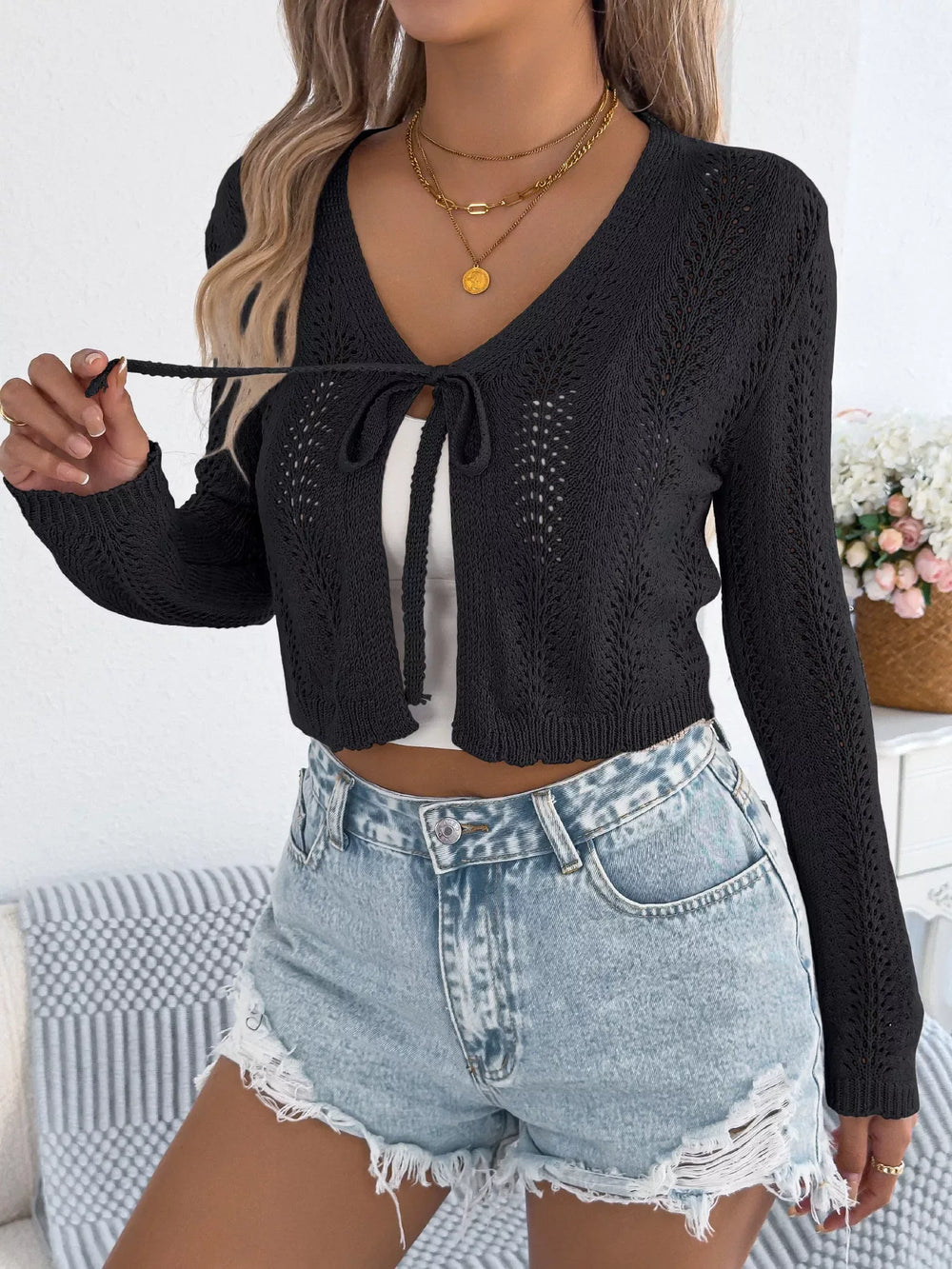 Woman wearing a black cropped knit cardigan with a tie front closure and denim shorts, adjusting the tie.
