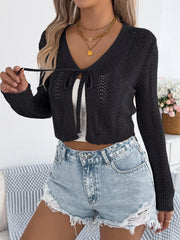 Woman wearing a black cropped knit cardigan with a tie front closure and denim shorts, adjusting the tie.
