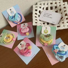 A collection of scrunchie sets in various pastel colors including pink, yellow, green, and blue, each with a cute charm, arranged on a wooden surface.