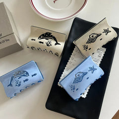 Collection of four hair claw clips with ocean-themed designs, including dolphins and seashells, in blue and cream colors