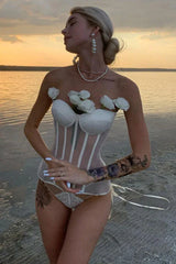 Woman wearing a white strapless boned corset with sheer panels and floral embellishments on a beach.