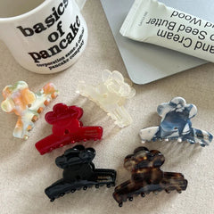 A collection of flower-shaped claw clips in various colors and patterns, including red, black, tortoiseshell, marbled blue and white, marbled orange and green, and translucent white, arranged on a textured surface next to a mug and a tube of hand cream.
