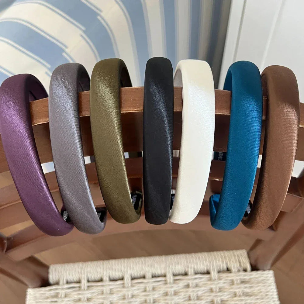 Collection of padded satin headbands in various colors (purple, grey, olive green, black, white, teal, brown) displayed on a wooden rack