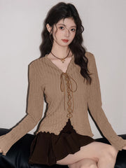 Woman sitting and wearing a tan ribbed knit long-sleeve top with a tie-front closure and ruffled hem, paired with a brown pleated skirt.