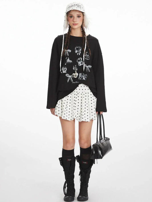 Full length studio shot of a young woman wearing a black long-sleeved graphic t-shirt, a white polka dot mini skirt, black knee-high socks, and black boots. She is also wearing a white polka dot bucket hat with fur trim and carrying a black handbag.