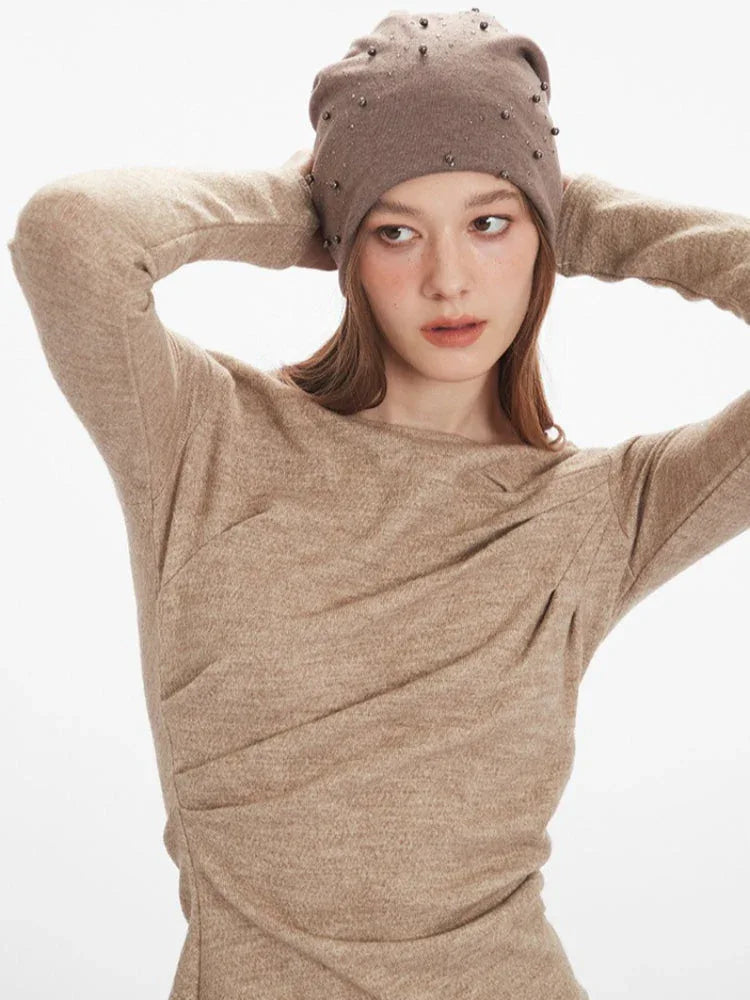 Close-up of a woman adjusting her beige beanie with studs while wearing a beige long-sleeve knit top.