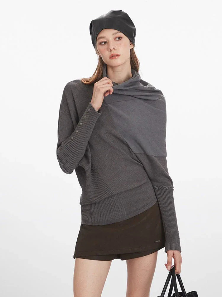 Close-up of a woman wearing a grey asymmetrical knit sweater with a draped neckline, brown shorts, and a black leather beret.