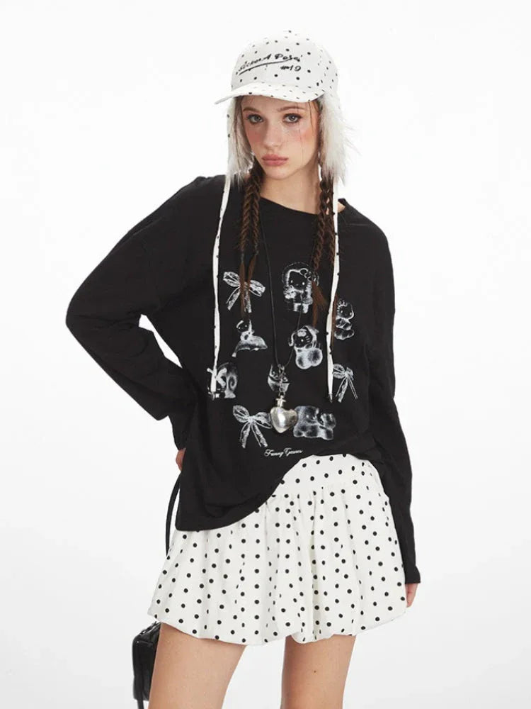 Front close-up of a young woman wearing a black long-sleeved graphic t-shirt with various charms printed on it. She is also wearing a white polka dot mini skirt and a white polka dot bucket hat with fur trim.