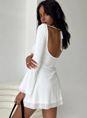 Back view of a woman wearing a white long-sleeve mini dress with a scoop back and tie detail.