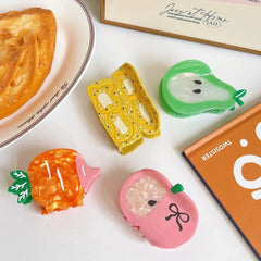 A collection of fruit-themed hair clips including a carrot, pear, banana slices, and an apple, arranged on a white surface near a pastry and a book.