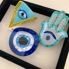 A collection of three evil eye themed accessories: a triangle, a round disc, and a Hamsa hand.