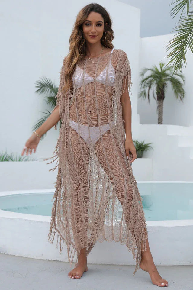 Woman wearing a beige open knit long tunic with fringe detailing and a side slit, standing by a pool.