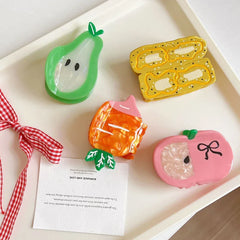 A white tray displaying fruit-shaped hair clips: a green pear, a yellow banana slice design, an orange carrot, and a pink apple, with a red and white checkered ribbon and a note.
