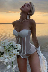 Woman wearing a white strapless boned corset with sheer panels, pearl jewelry, and a bouquet of white flowers.