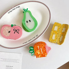 A white oval plate with text 'Mon cher Paris' holds a green pear hair clip and a pink apple hair clip. Next to it are a yellow banana slice hair clip and an orange carrot hair clip.