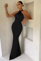 Woman leaning against a wall in a floor-length black satin halter dress, with one hand on the wall