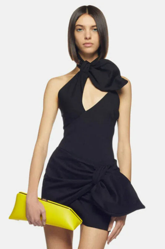 Woman wearing a black mini dress with a large bow detail at the halter neckline and a knot detail at the hem, holding a yellow clutch.