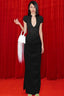 Model wearing a black long dress with a deep V-neckline and ruching on the skirt, holding a white bag with fringe details, standing in front of a red curtain.