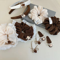 A variety of hair accessories, including scrunchies and hair clips, arranged on a surface with books. The accessories are in brown and cream, featuring lace and floral embellishments.