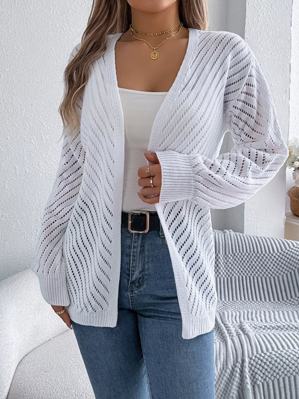 Front view of a woman wearing a white open-knit cardigan, white tank top, and blue jeans, with gold jewelry.