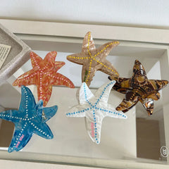 Collection of five starfish hair clips in various colors (orange, yellow, brown, blue, white) arranged on a glass surface