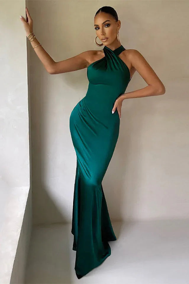 Woman leaning against a wall in a floor-length emerald green satin halter dress, with hands on hips