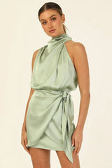 woman wearing a sage green satin halter neck mini dress with a draped bodice and tie waist
