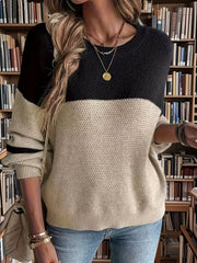 Front view of a woman in a black and tan color-blocked sweater with striped sleeves, layered necklaces, and blue jeans, with a bookshelf background.
