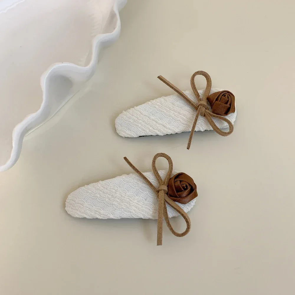 A pair of white fabric hair clips, each decorated with a brown rose and a thin brown string tied into a bow, presented on a neutral background.