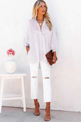 Full body shot of a woman wearing a white and pink pinstripe oversized button-down shirt, distressed white jeans, and tan heeled sandals, holding a brown clutch.