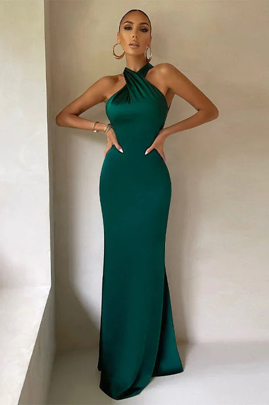 Woman wearing a floor-length emerald green satin halter dress with an open back, posing with hands on hips