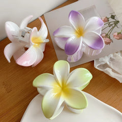 A selection of plumeria flower hair clips in pink, purple, and green, displayed on a white dish and a wooden surface.