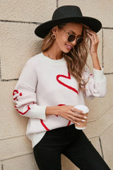 Woman wearing a white knit sweater with a red heart print, black hat, sunglasses, and holding a coffee cup, touching her hair.