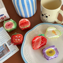 A top-down view of various fruit-shaped hair clips displayed on a table next to a mug and some packaging.
