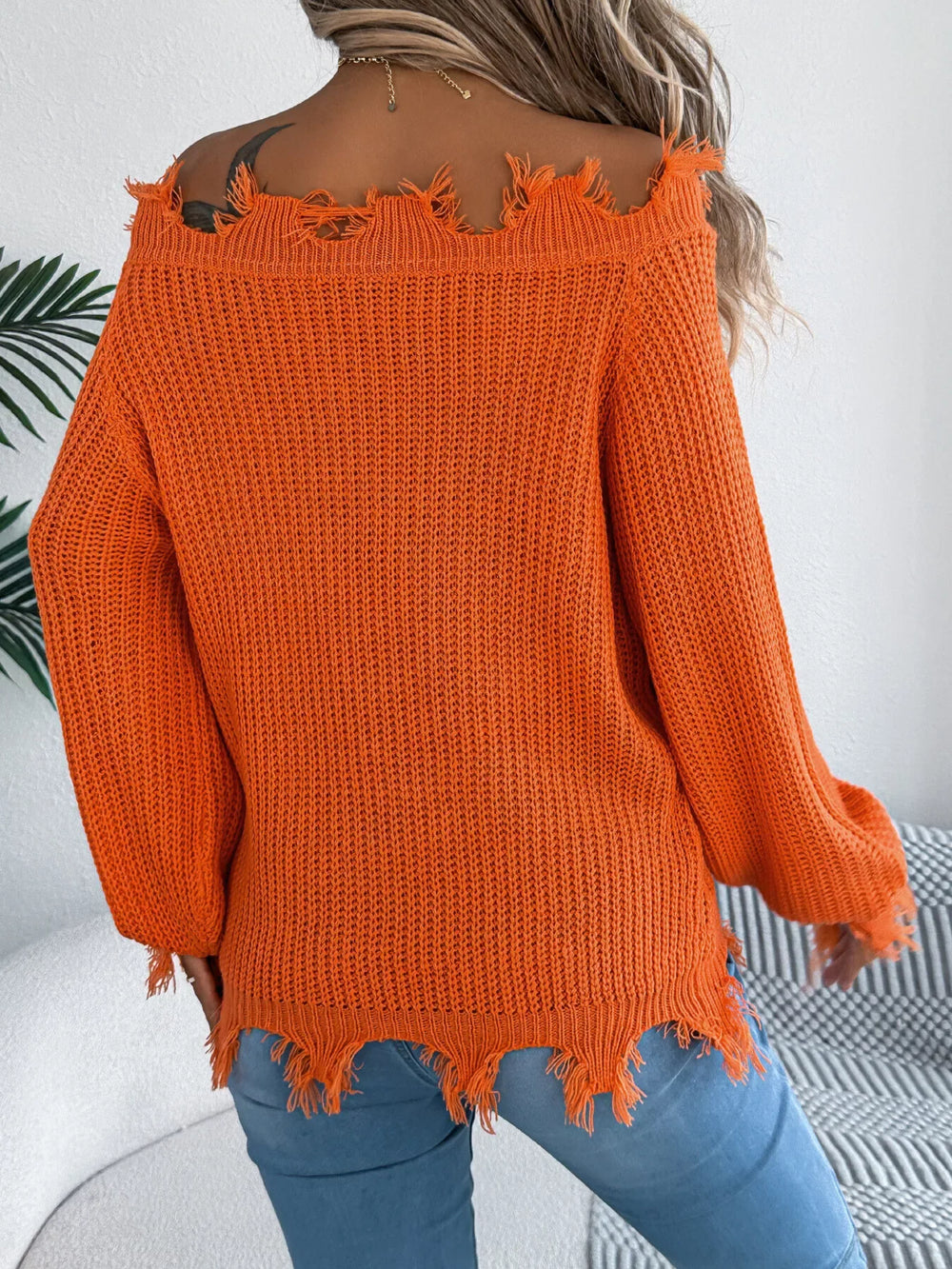Back view of a woman wearing an orange distressed off-the-shoulder knit sweater with blue jeans.