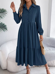 Woman standing holding a straw bag and wearing a long-sleeved blue midi dress with a collared neckline and tiered hem