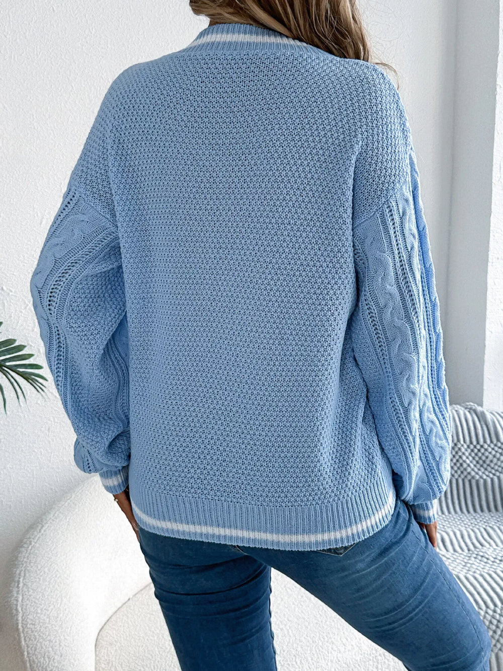 back view of a woman wearing a light blue cable knit v-neck sweater with white stripes and blue jeans