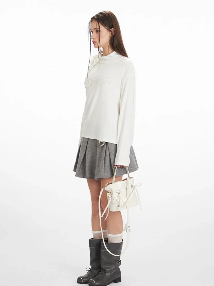 Side view of a woman wearing a white long-sleeved top with one shoulder exposed and a grey pleated skirt, holding a white handbag.