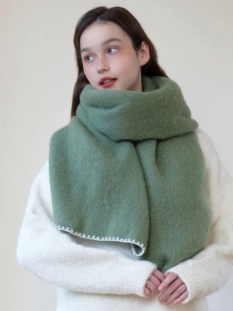 Woman wearing an oversized green scarf with a white fringed edge and a cream-colored sweater, looking to the side.