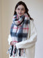 Woman wearing a cream-colored sweater and a plaid scarf, holding the ends of the scarf.