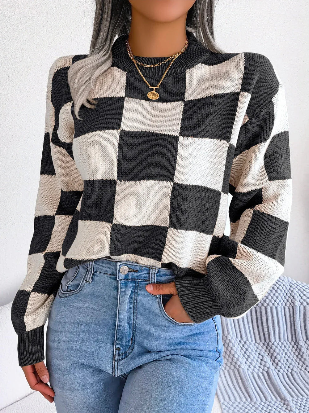 Woman wearing a black and white checkerboard knit sweater with light wash jeans and a gold necklace, front view.