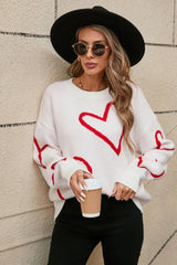 Woman wearing a white knit sweater with a red heart print, black hat, sunglasses, and holding a coffee cup, leaning against a brick wall.